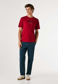 Raudona medvilninė marškinėlius su tamsiai mėlynu "Pepe Jeans London" logotipu. Suderinti su mėlynomis džinsinėmis kelnėmis ir baltais sportbačiais su rudu akcentu.