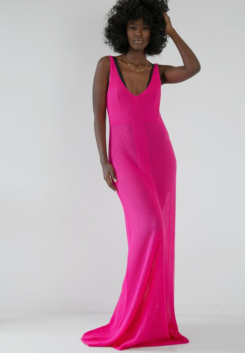Długa, intensywnie różowa sukienka z głębokim dekoltem w kształcie litery V i otwartą siateczkową strukturą. Posiada szerokie ramiączka oraz dopasowaną górę, która swobodnie opada do podłogi.