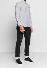 Šedá košile s límečkem, kapsou a dlouhými rukávy, doplněná černými slim-fit kalhotami a černými teniskami, se bílými ponožkami.