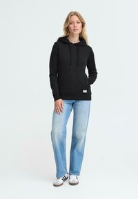 Felpa nera con cappuccio, dotata di tasca frontale e stringhe, abbinata a jeans azzurri chiari e sneakers bianche con strisce nere.
