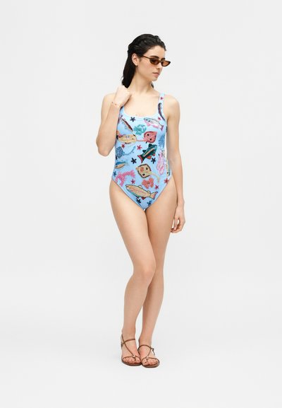 Γυναίκα που φοράει ολόσωμο μαγιό ανοιχτού γαλάζιου με πολύχρωμα σχέδια θαλάσσιων ζώων, καφέ σανδάλια και καφέ γυαλιά ηλίου, στέκεται μπροστά σε λευκό φόντο.