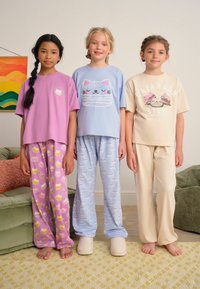 Tre bambini indossano completi di pigiami colorati: una maglietta viola con una stampa di torta, una maglietta blu con un disegno di gatto e una maglietta crema. I pantaloni hanno motivi giocosi.