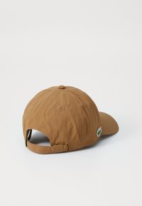 Casquette en coton brun avec une visière incurvée, une sangle ajustable et un patch logo sur le côté. Elle dispose d'œillets de ventilation et d'une finition texturée.