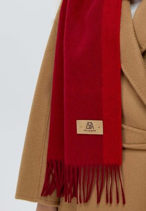 Roter Wollschal mit fransigen Enden, in rechteckiger Form. An ihm befestigt ist ein Lederschild mit der Prägung "BELLEMERE". Er wird über einem beigen Mantel getragen.