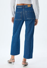 Jeans de pierna ancha de denim azul con textura desgastada, que presentan dos bolsillos traseros y un dobladillo sin acabar, combinados con sandalias de tacón plateadas.