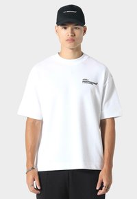 Witte oversized t-shirt met korte mouwen, voorzien van zwarte grafische tekst op de borst. Gecombineerd met een zwarte pet, met een neutrale achtergrond.