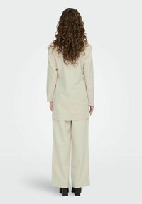 Beige skräddarsydd kostym med en lång blazer och vida byxor. Tyget verkar smidigt med en strukturerad silhuett och minimala detaljer.