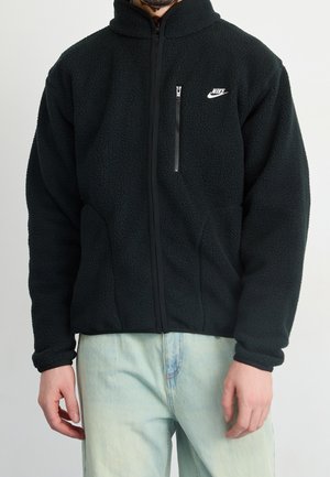 Homme portant une veste polaire noire Nike avec une fermeture éclair intégrale et une poche poitrine, associée à un jean denim bleu clair.