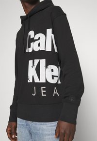 Felpa nera con grande scritta bianca "Calvin Klein JEANS." Presenta un cappuccio con cordini e un logo sul maniche. Tessuto morbido.