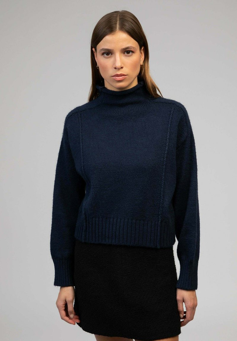 Pull en col roulé bleu marine avec un tissu texturé, des poignets et un ourlet côtelés, associé à une mini-jupe noire. Design simple, apparence chaleureuse.