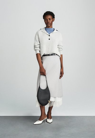 Világosszürke bordázott kötött pulóver fekete gombokkal, világosszürke térd hosszúságú szoknyával, fehér magassarkú mules cipővel és fekete táskával párosítva.