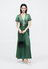 Rochie maxi din satin verde cu mâneci evazate și corset răsucit decorat cu un accesoriu argintiu. Asortată cu o geantă neagră mică și pantofi albi.