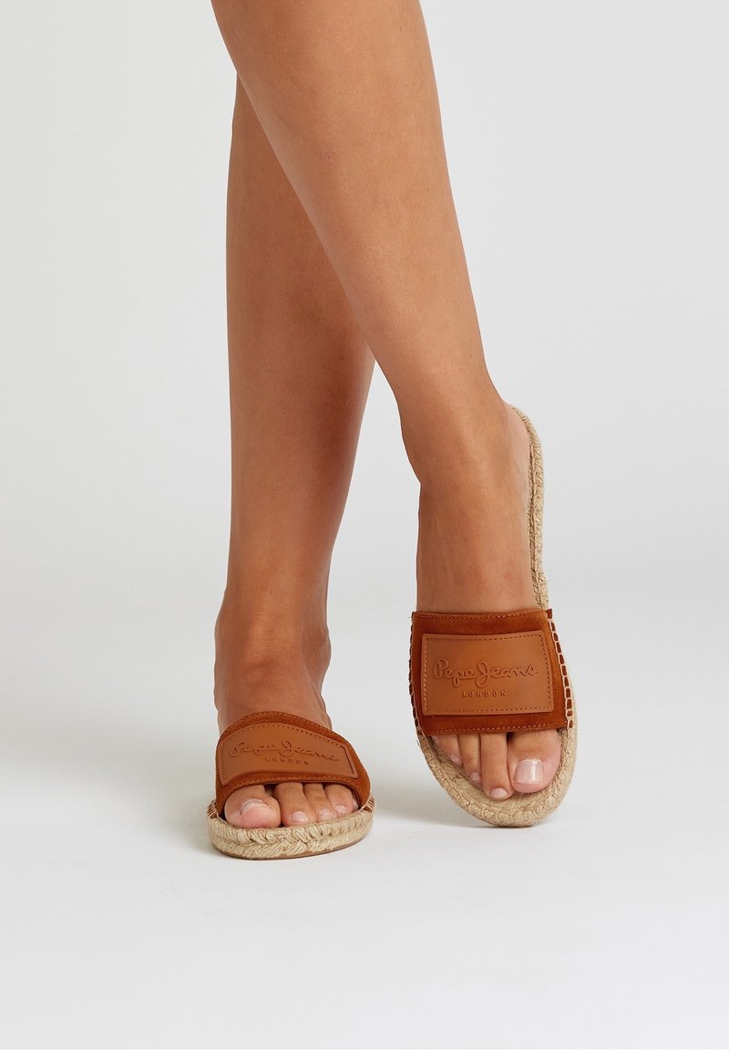 Pieds portant des sandales espadrilles marron à enfiler avec des semelles tissées et des brides embossées "Pepe Jeans London" sur un fond clair uni.