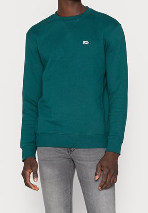 Mężczyzna w turkusowej bluzie crewneck z małym logo "Lee" na piersi oraz w jasnoszarych dżinsach na jednolitym tle.