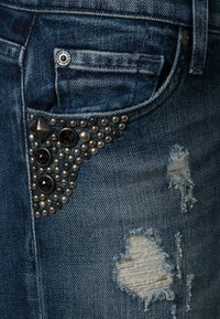 Vaqueros de mezclilla con textura desgastada, color azul oscuro y bolsillo frontal adornado con tachuelas negras y cuentas plateadas en un patrón triangular.