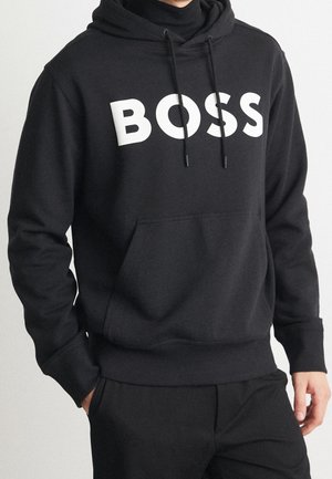 Mężczyzna noszący czarną bluzę z kapturem z przednią kieszenią i napisem "BOSS" dużymi białymi literami na piersi.