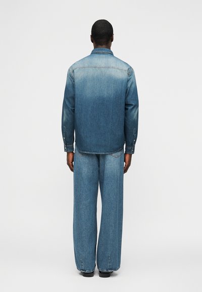 Άνδρας που στέκεται με την πλάτη, φορώντας ξεθωριασμένο μπλε τζιν μπουφάν και φαρδιά μπλε τζιν παντελόνια, με φόντο λευκό.