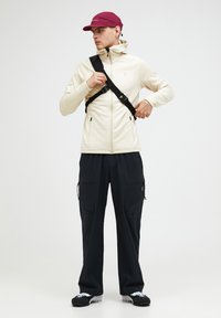 Beež zip-up jakk, kõrge kaelusega, mustad cargo-püksid ja marooni tooni müts. Must rihm üle rinna, tekstureeritud kangas ja tõmbelukuga taskud.