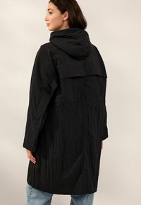 Manteau noir à capuche avec un tissu texturé, des manches longues et une coupe au genou. La vue arrière montre un détail arrière superposé et des boutons-pression.