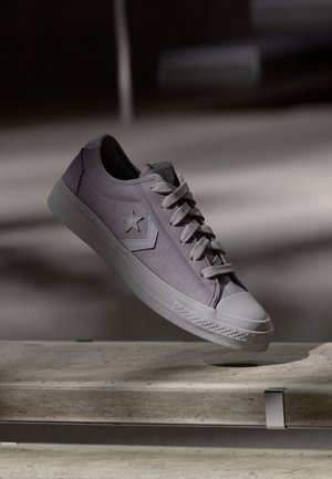 Shoe low-top em camurça lavanda clara com sola de borracha. Apresenta um logotipo em forma de estrela, atacadores brancos e uma biqueira texturizada. Exposição suspensa.