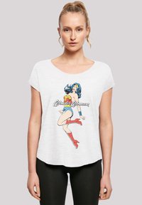 Valkoinen lyhythihainen T-paita, jossa on värikäs Wonder Woman -kuvio etupuolella, yhdistetty mustiin legginsseihin. Pehmeä, tekstuurilla varustettu kangas.