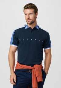Hombre con camisa polo azul marino con detalles azules y texto blanco "HACKETT", sudadera color óxido atada alrededor de la cintura, manos en los bolsillos.