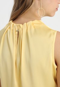 Blusa amarilla sin mangas hecha de una tela suave y ligera. Presenta un escote fruncido y un pequeño cierre de botón en la parte posterior.