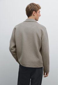 Beige kabát z vlněné směsi se strukturovaným límcem, jednoduchým designem a mírně oversized střihem. Má čistou, hladkou texturu a minimální detaily.