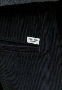 Tissu en velours côtelé noir avec des côtes verticales, présentant une étiquette blanche portant l'inscription "JACK & JONES EST. 1990" attachée à une poche.