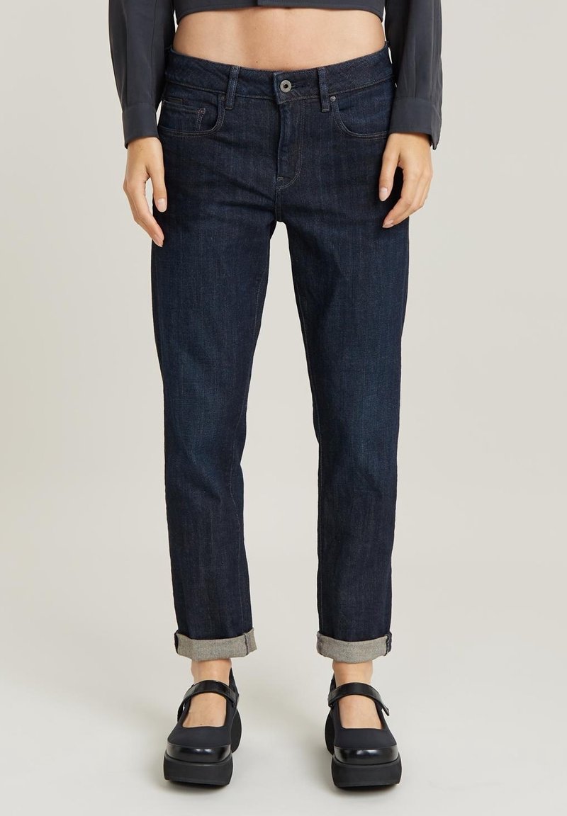 Tummansiniset farkut, joissa on suora lahje, käännetyt lahkeensuuhat ja perinteinen napitus. Huomaamattomat tikkausyksityiskohdat näkyvissä.