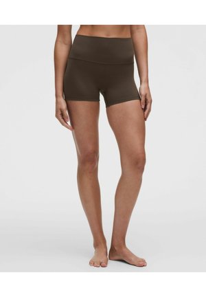 Kvinde iført højtaljede, mørkebrune, tætsiddende sportsshorts, stående barfodet mod en ensfarvet grå baggrund.