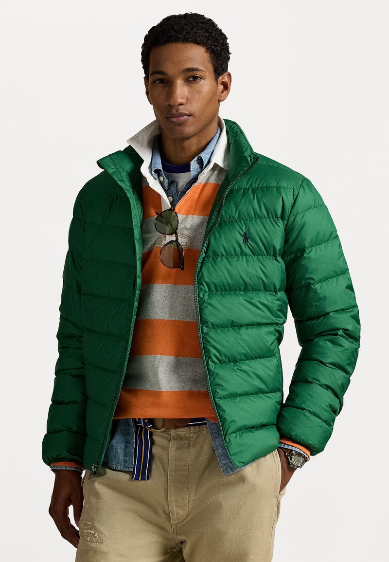 Grønne pufferjakke med quiltet design, iført over en grå og orange stribet sweater, lagdelt med en denimskjorte, beige bukser synlige.