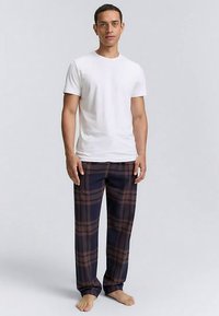 Weißes T-Shirt mit kurzen Ärmeln, kombiniert mit marineblauen Karotten-Pyjama-Hosen mit braunen Linien. Mann steht barfuß vor einem einfarbigen Hintergrund.