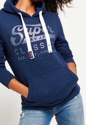 Personne portant un sweat à capuche bleu Superdry avec des cordons blancs et une poche avant, associé à un jean bleu clair déchiré.