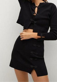 Pull court noir avec des fermetures décoratives à nœuds, associé à une jupe noire ajustée présentant des détails de nœuds similaires et un ourlet légèrement asymétrique.