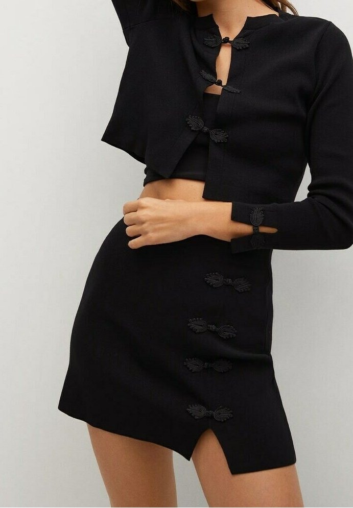 Pull court noir avec des fermetures décoratives à nœuds, associé à une jupe noire ajustée présentant des détails de nœuds similaires et un ourlet légèrement asymétrique.