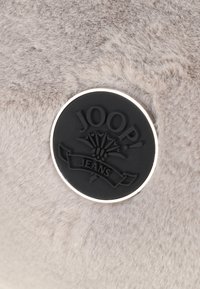 Tessuto in pelliccia sintetica grigia con un logo rotondo in gomma nera che riporta "JOOP! JEANS" e un design floreale, circondato da una bordatura in argento.