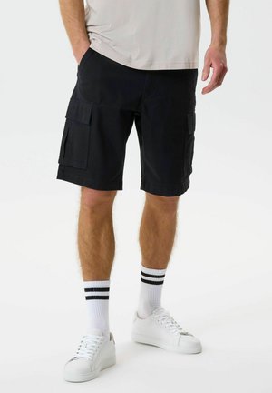 Férfi fekete cargo rövidnadrágban, fehér zokniban fekete csíkokkal, fehér sportcipőben és világos színű pólóban, fehér háttér előtt áll.