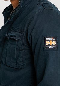 Marinblå militärjacka med dragkedja och tryckknappsstängning, med en logotyp-patch på vänster ärm och en ficka framtill.