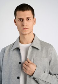 Ljusgrå kragjacka med knappstängning, bröstficka och slät textur, buren över en vit t-shirt med rund hals.