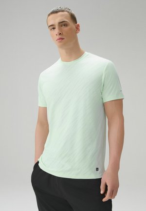 Junger Mann mit kurzem Haar, der ein hellmintgrünes kurzärmeliges T-Shirt und schwarze Hosen trägt und mit einer Hand in der Tasche steht.