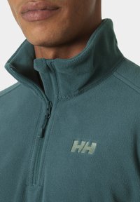 Turkusowy polarowy pulower z wysokim kołnierzem, zamkiem błyskawicznym na ćwierć długości i wytłoczonym logo "HH" w jaśniejszym odcieniu. Miękka, teksturowana tkanina.