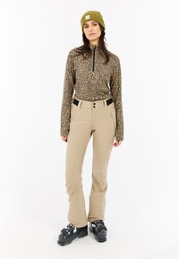 Top con estampado de leopardo en tonos marrones y negros, combinado con pantalones beiges con frente plano y bolsillos laterales con cremallera. Botas de esquí negras.