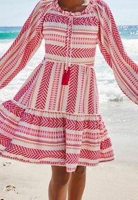 Vestido a rayas rojas y blancas con mangas largas, que presenta un dobladillo con volantes, detalles de flecos y patrones geométricos en la tela.