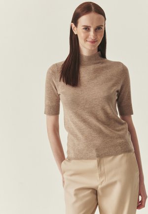 Kobieta o długich brązowych włosach ubrana w beżowy sweter z krótkim rękawem i jasno-brązowe spodnie, stoi z jedną ręką w kieszeni, delikatnie się uśmiechając.