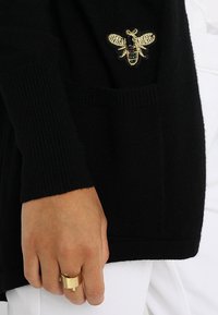 Svart stickad kofta med en guldfärgad och svart broderad bi på fickan. Den visade handen bär en guldring, med vit bakgrund.