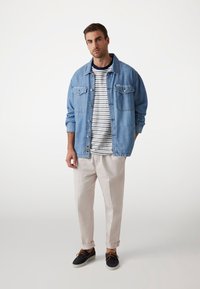 Chaqueta de mezclilla en azul claro, diseño de botones con dos bolsillos en el pecho, llevada sobre una camiseta de rayas blanca y azul marino, combinada con pantalones claros y zapatos.