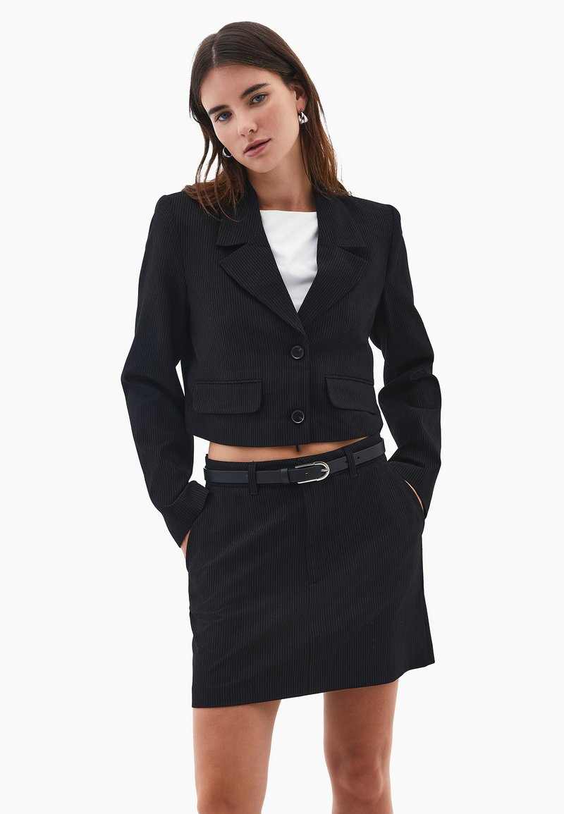 Chaqueta corta negra a rayas con dos bolsillos frontales, cierre de un botón y falda a juego, combinada con un cinturón negro y una blusa blanca.