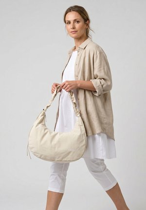 Kvinde iført hvid kjole og beige skjorte, der holder en stor beige håndtaske med samlet håndtag og "Venezia"-logo.