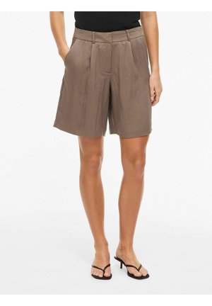 Naine, kes kannab lahtisi, pruunid põlvikupikkused lühikesed püksid volditud detailidega, musta t-särki ja musti kontsaga sandaale.